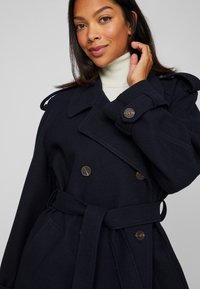 Γυναίκα φορώντας ναυτική κολόνια παλτό με ζώνη πάνω από λευκό καστόνι, αγγίζοντας τα μαλλιά της, με ουδέτερη έκφραση σε ανοιχτό γκρι φόντο.