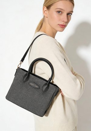 Femme blonde en pull crème portant un sac à main noir texturé avec double poignée et bandoulière, étiqueté « pierre cardin ».
