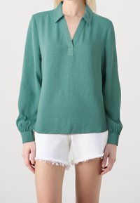 Blusa verde con colletto e uno spacco frontale. Caratterizzata da maniche lunghe e puffate con dettagli ai polsini. Abbinata a pantaloni corti bianchi sfilacciati.