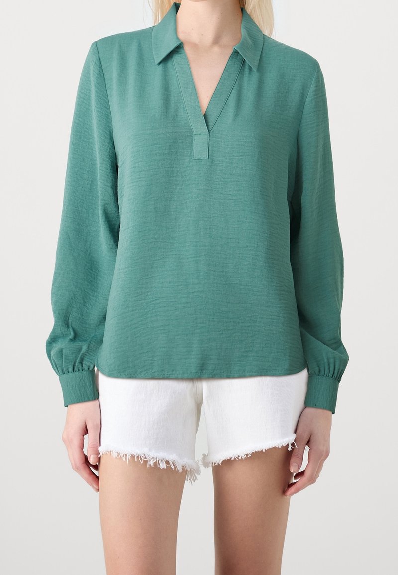 Blusa verde con colletto e uno spacco frontale. Caratterizzata da maniche lunghe e puffate con dettagli ai polsini. Abbinata a pantaloni corti bianchi sfilacciati.