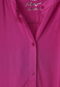 Camicia fucsia con colletto, caratterizzata da una leggera lucentezza, chiusura con un singolo bottone e dettaglio plissettato sul retro.