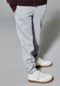 Grijze sweatpants van zacht materiaal, met een relaxte pasvorm en geribde boorden, gecombineerd met witte sneakers met een klittenbandsluiting en een bruine buitenzool.