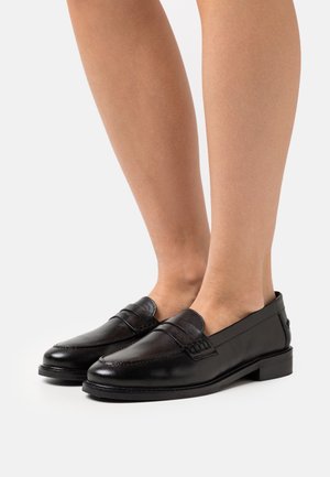 Mocassins en cuir noir avec une finition lisse, un faible talon, des coutures surélevées et une sangle sur le devant. Un design à la fois élégant et fonctionnel.