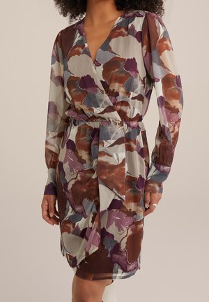 Femme portant une robe portefeuille à manches longues avec un motif floral abstrait dans des nuances de marron, violet, gris et blanc.