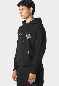 Černá mikina s kapucí a přední kapsou, zdobená nášivkou "PAS DE MONACO" a kostkovanými akcenty. Měkká tkanina, žebrované manžety a šňůrky.