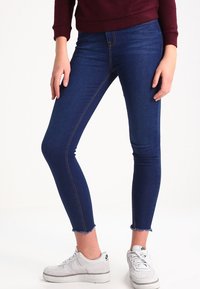 Vaqueros ajustados de mezclilla azul oscuro con dobladillo deshilachado, con costuras contrastantes y un ajuste ceñido. Combinados con zapatillas blancas.