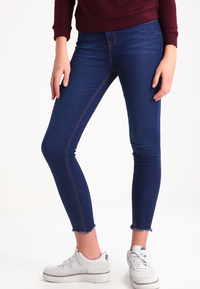 Vaqueros ajustados de mezclilla azul oscuro con dobladillo deshilachado, con costuras contrastantes y un ajuste ceñido. Combinados con zapatillas blancas.