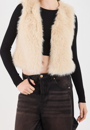 Femme portant un gilet sans manches en fausse fourrure beige sur un crop top noir côtelé à manches longues et un jean foncé taille haute.
