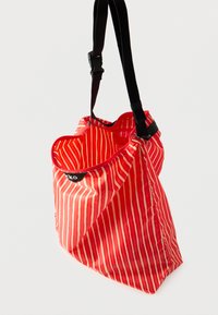 Τσάντα ώμου με κόκκινες και λευκές ρίγες, κατασκευασμένη από ελαφρύ, γυαλιστερό υλικό. Διαθέτει μαύρο ρυθμιζόμενο λουρί και φερμουάρ στην κορυφή.