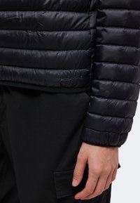 Gros plan de la main d'une personne près de la manche d'une veste matelassée noire Patagonia et d'un pantalon noir avec une poche latérale.