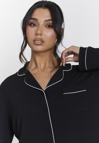 Zwarte pyjama-top met witte bies, knoopsluiting, kraag en een zak op de linkerborst met een ribbelstructuur.