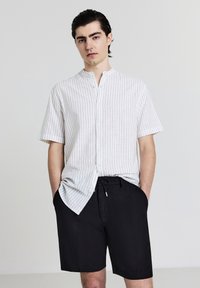 Kurzärmliges, knöpfbares Hemd in Weiß mit schwarzen senkrechten Streifen, kombiniert mit schwarzen Shorts mit Kordelzug und Seitentaschen.
