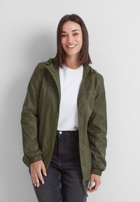 Smiling woman with straight dark hair wears an olive green zip-up jacket over a white t-shirt and black jeans against a white background.