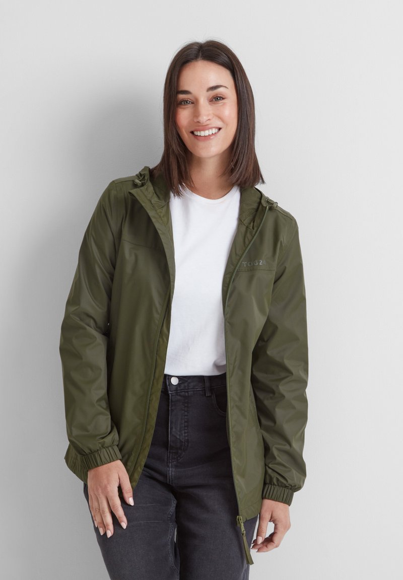 Smiling woman with straight dark hair wears an olive green zip-up jacket over a white t-shirt and black jeans against a white background.