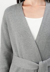 Szary dziergany kardigan z kołnierzem szalowym, z przodu z paskiem do związania i ściągaczami przy mankietach, noszony na białej bluzce z kwadratowym dekoltem.