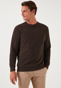 Pulover texturat de culoare maro, cu guler rotund și mâneci lungi, asortat cu pantaloni bej deschis. Design simplu, fără logo-uri sau modele vizibile.