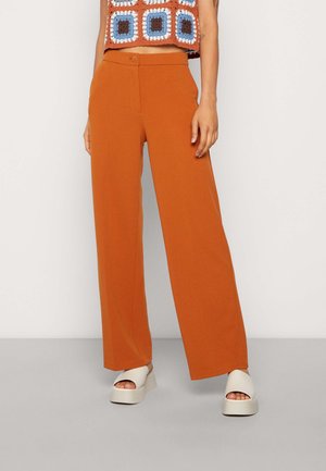 Model draagt wijde broek in gebrande oranje tint, witte platformsandalen en een cropped gehaakte top met vierkante patronen in blauw, wit en bruin.