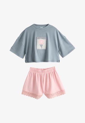 Smilende pige med brunt hår iført en løs blå skjorte med et blomsterpatch og lyserøde shorts med blonderkant mod en brun baggrund.
