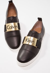 Chaussures slip-on en cuir noir avec une semelle blanche, ornées d'une bande métallique dorée portant le nom "Calvin Klein" sur des bandes élastiques. Texture lisse, bout arrondi.