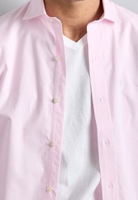 Světle růžová kostkovaná košile s knoflíky a bílým tričkem s výstřihem do V pod ním. Košile má jemnou texturu a řadu knoflíků.