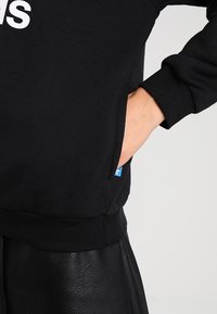 Svart sweatshirt med rundad halsringning, ribbade mudd i ärmar och nederkant. Har en sid ficka och en liten blå logotagg vid sömmen.