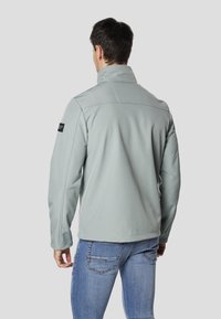 Jasnoszara kurtka softshell z kołnierzem stójką, przeszyciami i czarną naszywką z logo na lewym rękawie, noszona na niebieskich dżinsach.