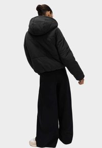 Giacca a vento nera corta con cappuccio, texture trapuntata e polsini elastici. Abbinata a pantaloni neri larghi e calzature leggere.