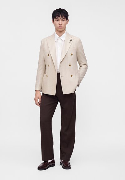 Jeune homme debout portant un blazer beige croisé avec des boutons dorés, une chemise blanche, un pantalon marron foncé et des chaussures marron foncé.