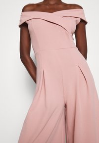 Femme portant une combinaison sans manches, décolletée aux épaules, de couleur rose pâle, avec des jambes larges et des plis à la taille.