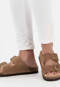 Sandali in suede marrone con doppia fibbia, soletta sagomata e suole testurizzate. Indossati con pantaloni bianchi aderenti, che rivelano le dita dei piedi e i dettagli del piede.