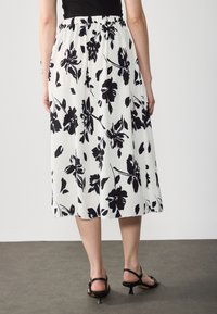 Jupe midi plissée blanche avec un grand motif floral noir. Ceinture élastique, texture fluide et coupe décontractée. Sandales noires à brides visibles.