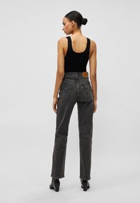 Γυναίκα που στέκεται με την πλάτη προς τα εμάς, φορώντας μαύρο αμάνικο τοπ, ψηλόμεσα ξεθωριασμένα μαύρα τζιν και μαύρα μποτάκια με τακούνι, μπροστά σε απλό φόντο.