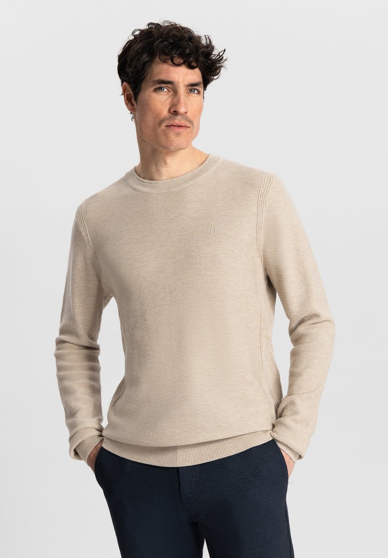 Beige strikket genser med rund hals, teksturert finish og ribbestrikkede mansjetter. Med logo på brystet. Kombinert med mørke bukser.
