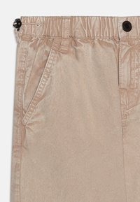 Pantalon beige en tissu lisse avec une taille élastique. Comprend des poches latérales, un accent de bouton et des détails de couture visibles.