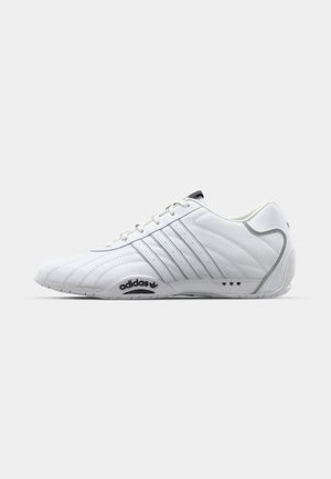 White athletic shoe with textured leather, lace-up design, three stripe accents, and black logo on the sole. Features rounded toe and low profile.
