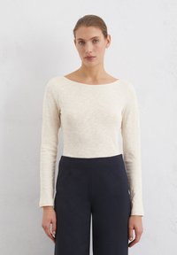 Helle beige Bluse mit langen Ärmeln und Bootsausschnitt, aus strukturiertem Stoff. Kombiniert mit dunkelblauen, weitgeschnittenen Hosen.