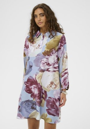 Kvietkové šaty s veľkými vzormi pivoniek v odtieňoch fialovej, krémovej a modrej, s golierovým výstrihom a dlhými, mierne nafúknutými rukávmi.