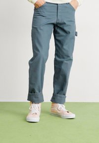 Blue cotton pants with rolled cuffs, featuring a side pocket. Paired with light pink and cream sneakers on a green surface.