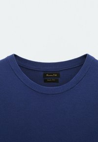 Granatowy, dzianinowy sweter z okrągłym, ribbowanym dekoltem. Tkanina ma teksturowany splot, a na metce znajduje się czarna etykieta z nazwą marki i szczegółami kolekcji.