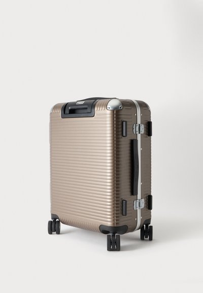 Valise rigide bronze debout avec quatre roues pivotantes noires et poignées noires sur fond blanc.