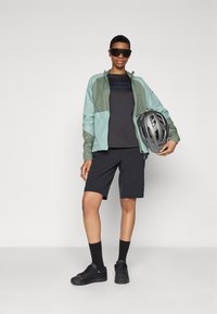 Atletický outfit s svetlozelenou a sivou bundou, čiernym tričkom, čiernymi šortkami a čiernymi teniskami. Drží sivú cyklistickú prilbu.