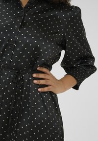 Zwarte satijnen jurk met crèmekleurige stippen, knoopsluiting aan de voorkant en een strik taille. Poffende mouwen met smock manchetten. Gladde textuur.