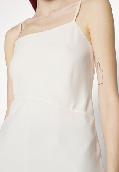 Personne portant une robe blanche sans manches avec de fines bretelles et un tatouage représentant le contour d'une église sur le haut de son bras.