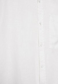 Détail d'une chemise en lin blanc montrant des boutons sur le devant et une poche poitrine côté droit.