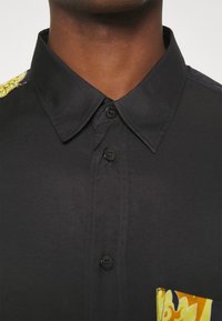 Zwarte korte mouwen shirt met een puntige kraag, voorzien van een gele bloemenzakdetail en vijf knopen aan de voorkant voor sluiting.