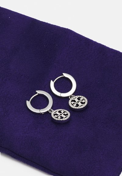Srebrne školjkaste naušnice s elementima koji vise, s crno-bijelim uzorkom, ukrašene prozirnim rhinestones, smještene na ljubičastoj tkanini.