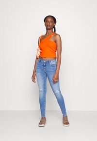 Top laranja de um ombro só em tecido canelado, combinado com jeans skinny azul claro com detalhes desgastados e sandálias planas bege.