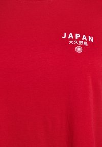 Sarkans kokvilnas T-krekls ar baltu drukājumā tekstu "JAPAN" un apakšā esošu apļveida ikonu ar japāņu rakstzīmēm. Gluda tekstūra.
