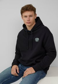 Zwarte hoodie met een voorzak, een trekkoordkap en het "Evolving Roots" logo in lichtblauw. Gemaakt in combinatie met blauwe jeans, met een soepele stoftextuur.