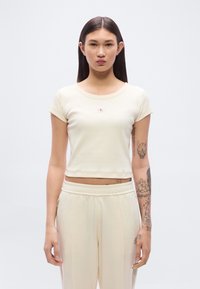 Ανοιχτό μπεζ κοντό t-shirt με κοντά μανίκια και στρογγυλή λαιμόκοψη, με ένα μικρό κόκκινο και λευκό λογότυπο στο στήθος.